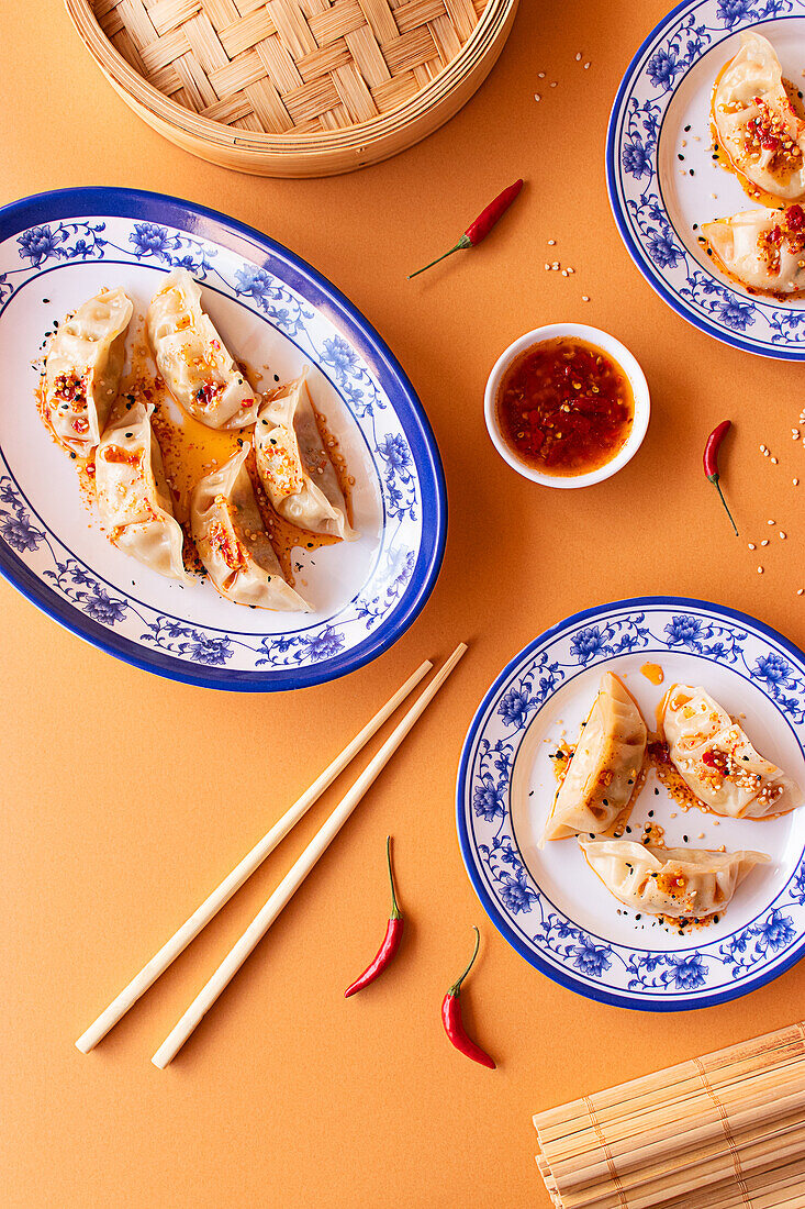 Spicy Chinese dumplings served with chili sauce in elegant blue-and-white patterned plates, complemented by rustic bamboo chopsticks and a bamboo steamer on a textured orange background