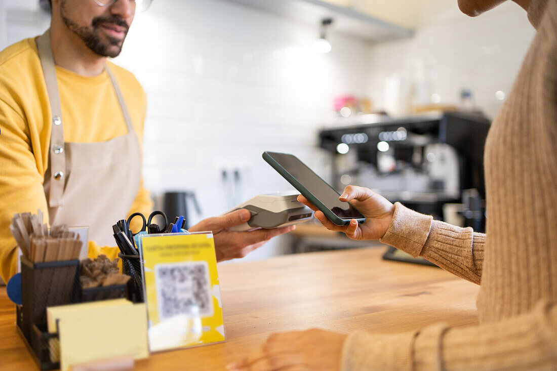 Ein Kunde benutzt sein Smartphone, um an der Kasse eines Cafés kontaktlos zu bezahlen, wobei er von einem freundlichen Barista unterstützt wird.