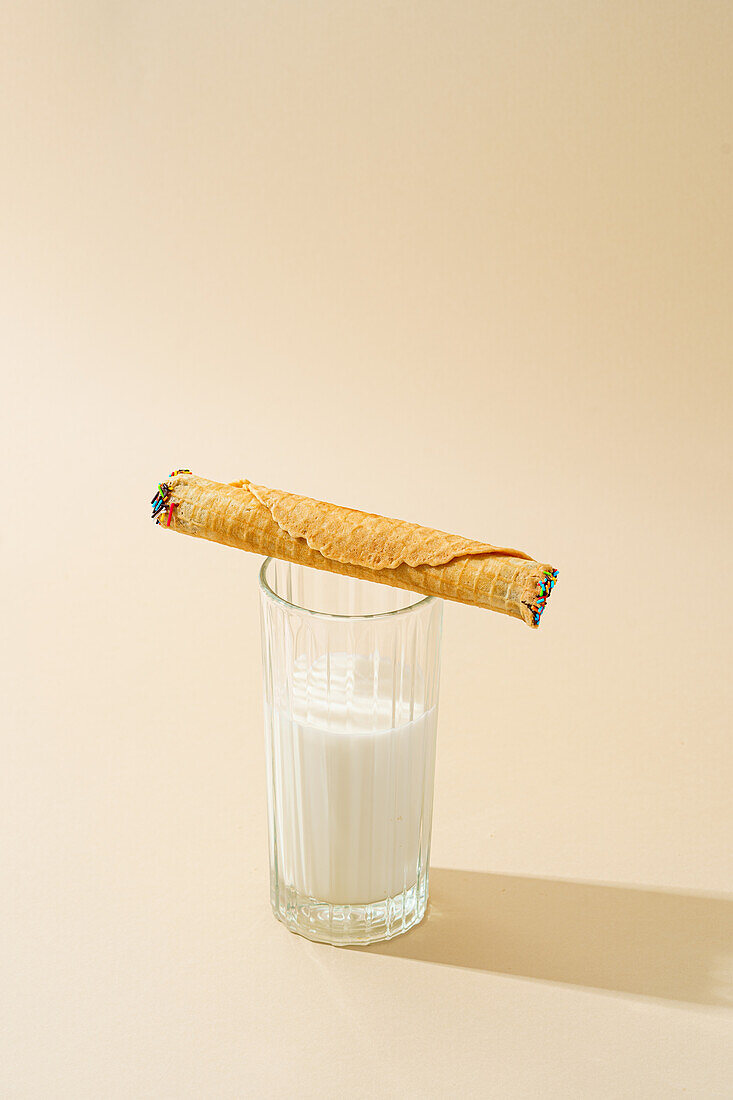 A crispy waffle roll filled with condensed milk leaning on a full glass of milk against a beige background.