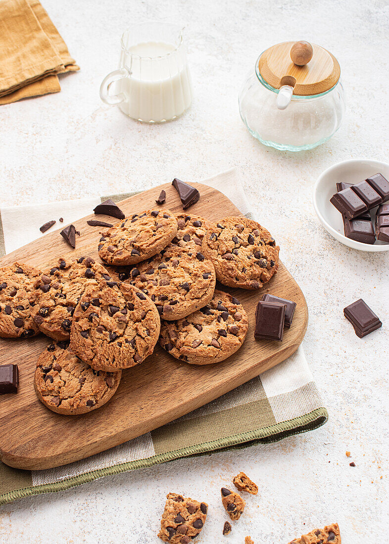 Eine köstliche Auswahl an Schokoladenkeksen auf einem Holzbrett, begleitet von Schokoladenstückchen, einem Milchglas und einer Zuckerdose im Hintergrund - perfekt für Dessert- und Snackkonzepte