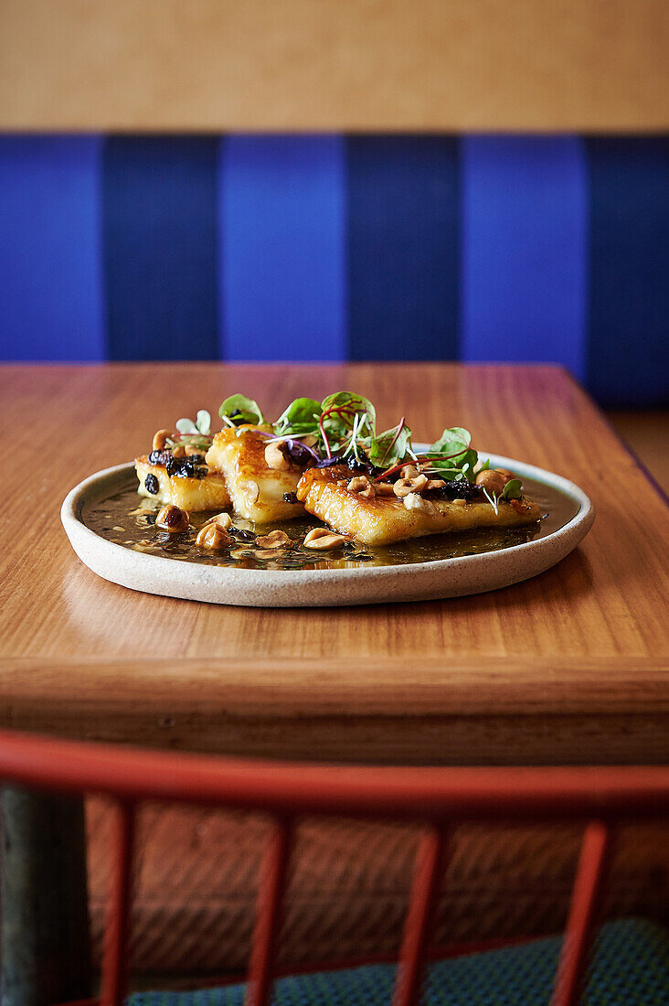 Grilled halloumi cheese served on a ceramic plate with seeds and microgreens, presented on a wooden table with a blue upholstered booth in the background.