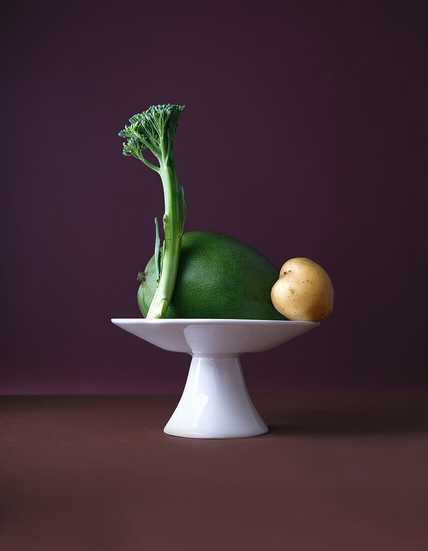 A creative display featuring a mango, potato, and broccoli stalk artfully arranged on a small, elegant white pedestal against a rich purple background