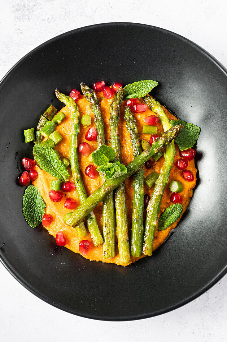 Blick von oben auf ein Gourmetgericht mit Süßkartoffelpüree, gegrilltem Spargel, frischen Granatapfelkernen und Minzblättern, serviert auf einem schwarzen Teller.