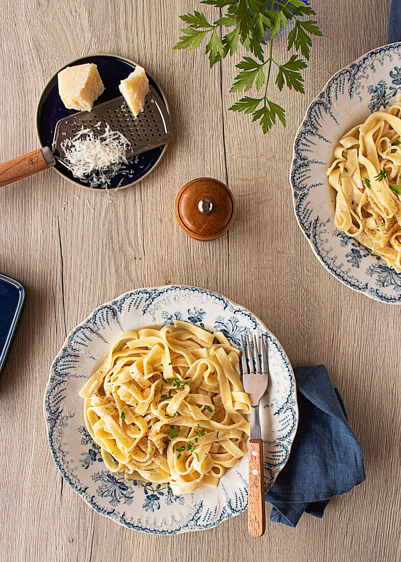 Draufsicht auf ein leckeres italienisches Nudelgericht mit cremiger Alfredo-Sauce, garniert mit Petersilie auf einem Holztisch mit Pfeffermühle und Parmesanreibe