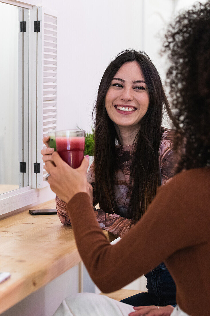 Content female sitting at counter drinking refreshing smoothie with cropped anonymous ethnic friend while chilling in bar and looking at each other