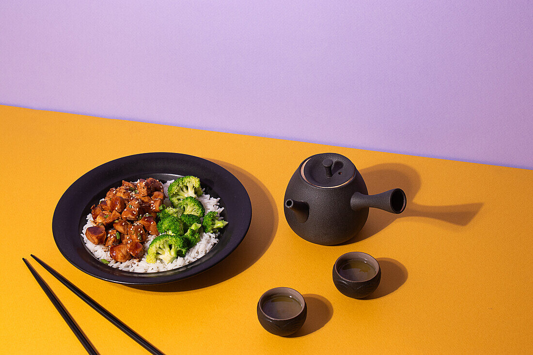 Kunstvoll angerichtetes Teriyaki-Hähnchen auf einem schwarzen Teller, serviert mit Brokkoli und Reis, begleitet von einem traditionellen Teeservice auf einem leuchtend gelben und violetten Hintergrund.