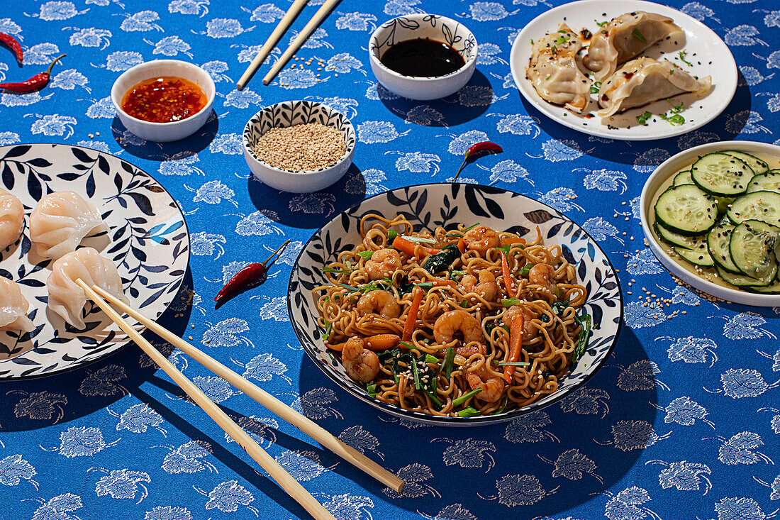 A vibrant assortment of Chinese food served on decorative dishes. Featured are noodle dish with shrimp, steamed dumplings, cucumber salad, and sauces