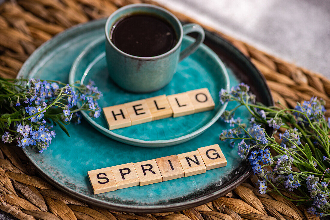 A cozy coffee setup with a ceramic mug on a teal plate, accompanied by vibrant forget-me-not flowers and welcoming HELLO SPRING spelled out in letter tiles