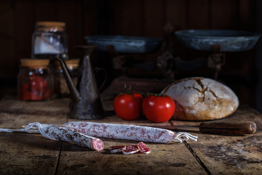 A vintage-styled kitchen table elegantly arranged with various cured meats, including sliced fuet with its distinctive red hue from the paprika, accompanied by fresh tomatoes, artisanal bread, and classical kitchenware.