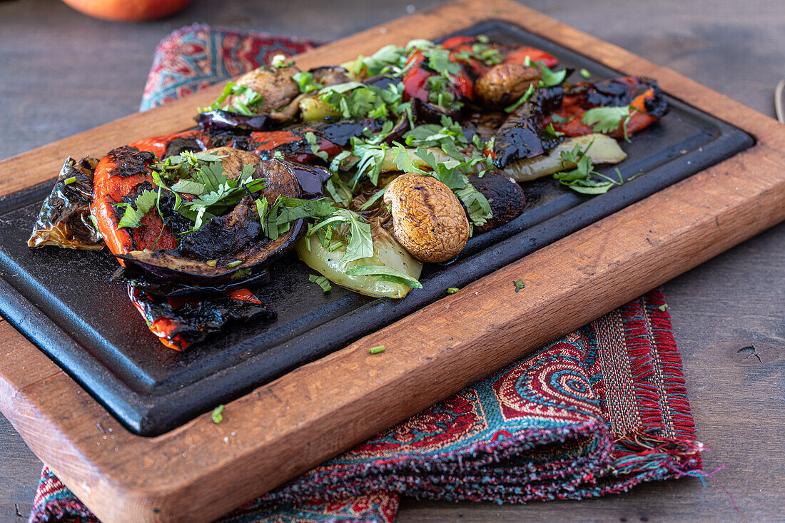 Colorful mix of grilled vegetables, including eggplants, peppers, and mushrooms, garnished with fresh herbs on a wooden serving board