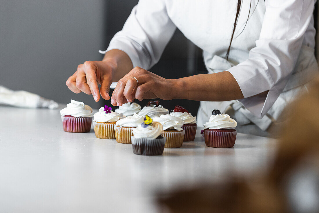 Anonyme Bäckerin dekoriert Cupcakes mit Blumen und Beeren, während sie in einer Bäckerei gesunde vegane Desserts zubereitet