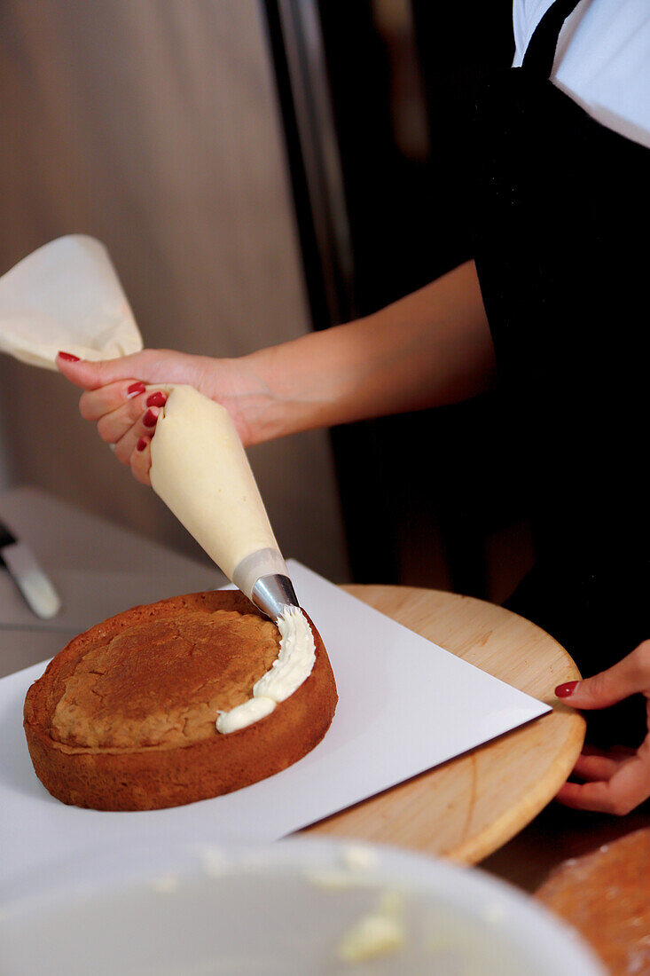 Close-up of a baker applying frosting on a cake with a piping bag in a kitchen setting, showcasing the art of cake decorating