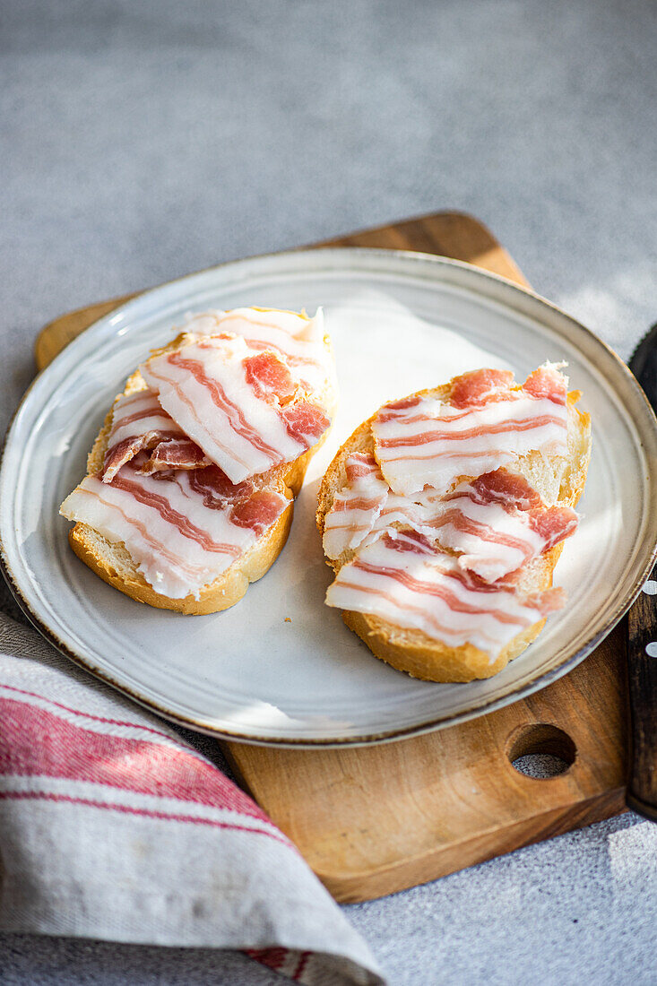 Zwei mit dünn geschnittenen Schmalzscheiben belegte Toasts, die auf einem Keramikteller mit Holzbrett und Stoffserviette präsentiert werden, von oben.