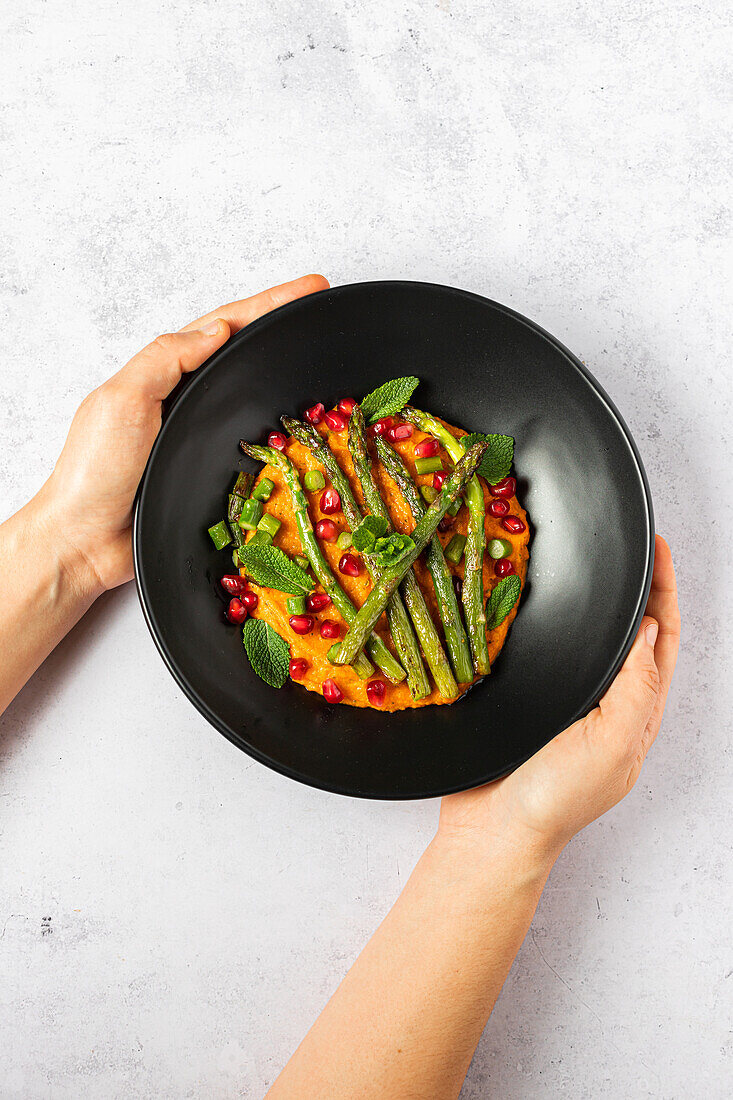 From above cropped unrecognizable hands holding a black plate of mashed sweet potato with mint, grilled asparagus, and pomegranate, top view.