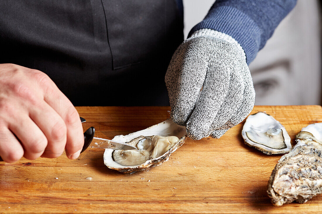 A chef's hands, wearing protective gloves, expertly opening fresh oysters with a specialized knife on a wooden board.