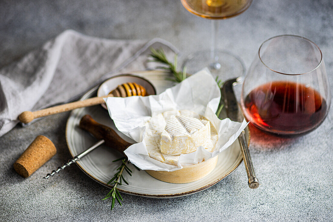 A sophisticated setup featuring Brie cheese on a plate, paired with a glass of red wine, complete with a honey dipper and rosemary.