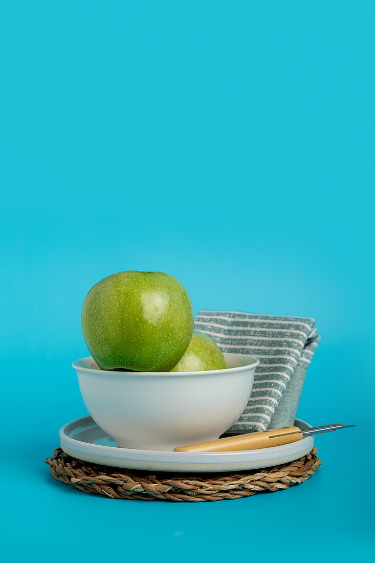 Crisp green apples nested elegantly in a white ceramic bowl, set above a rustic straw placemat on a striking turquoise background, complete with a casual striped napkin and a sleek knife for a touch of natural dining elegance.