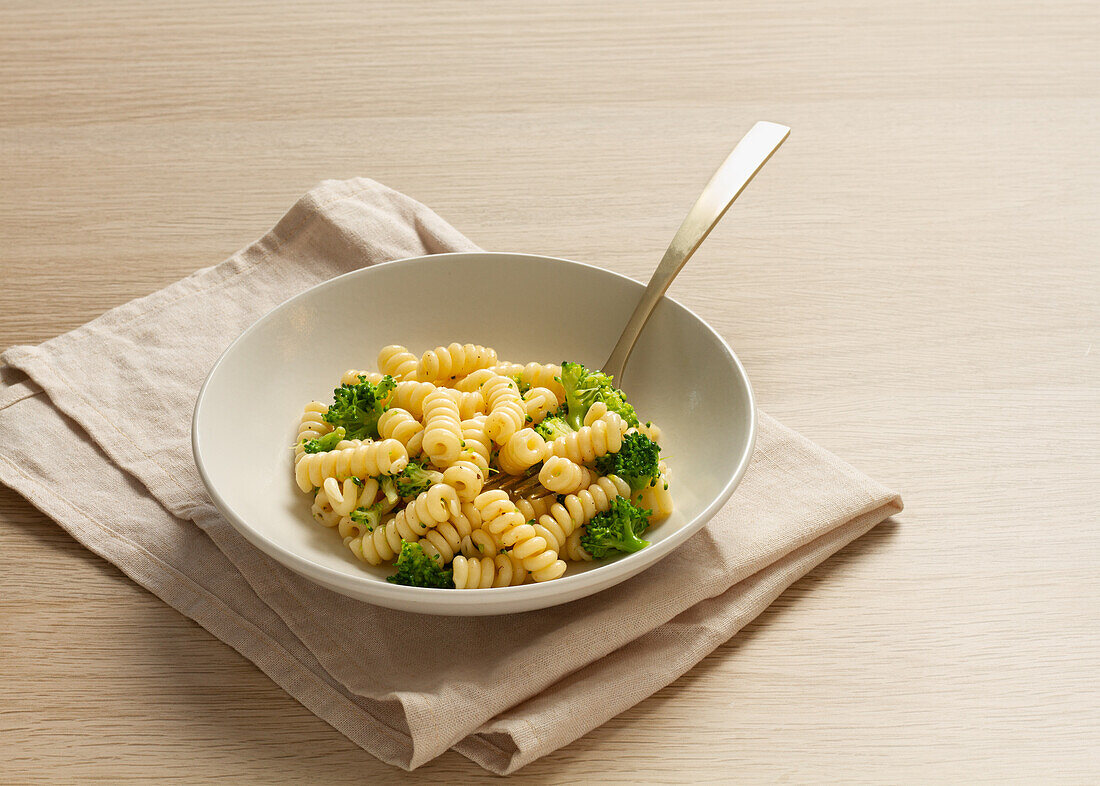 Eine ruhige Szene mit einer Schüssel Fusilli-Nudeln mit gedünstetem Brokkoli, begleitet von einer beigen Leinenserviette und einer modernen Gabel auf einer Holzoberfläche.