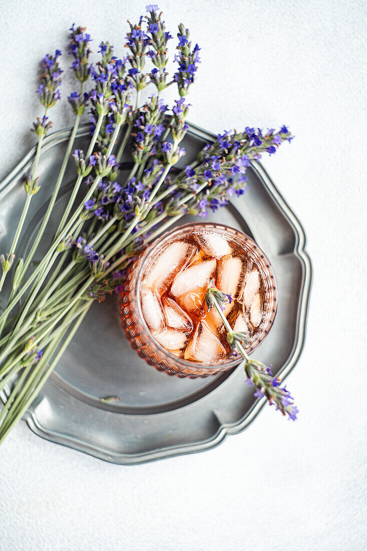 An elegant serving of a cognac cocktail enhanced with fresh lavender, showcased on a textured silver tray against a soft white background, offering a sophisticated, floral-themed libation