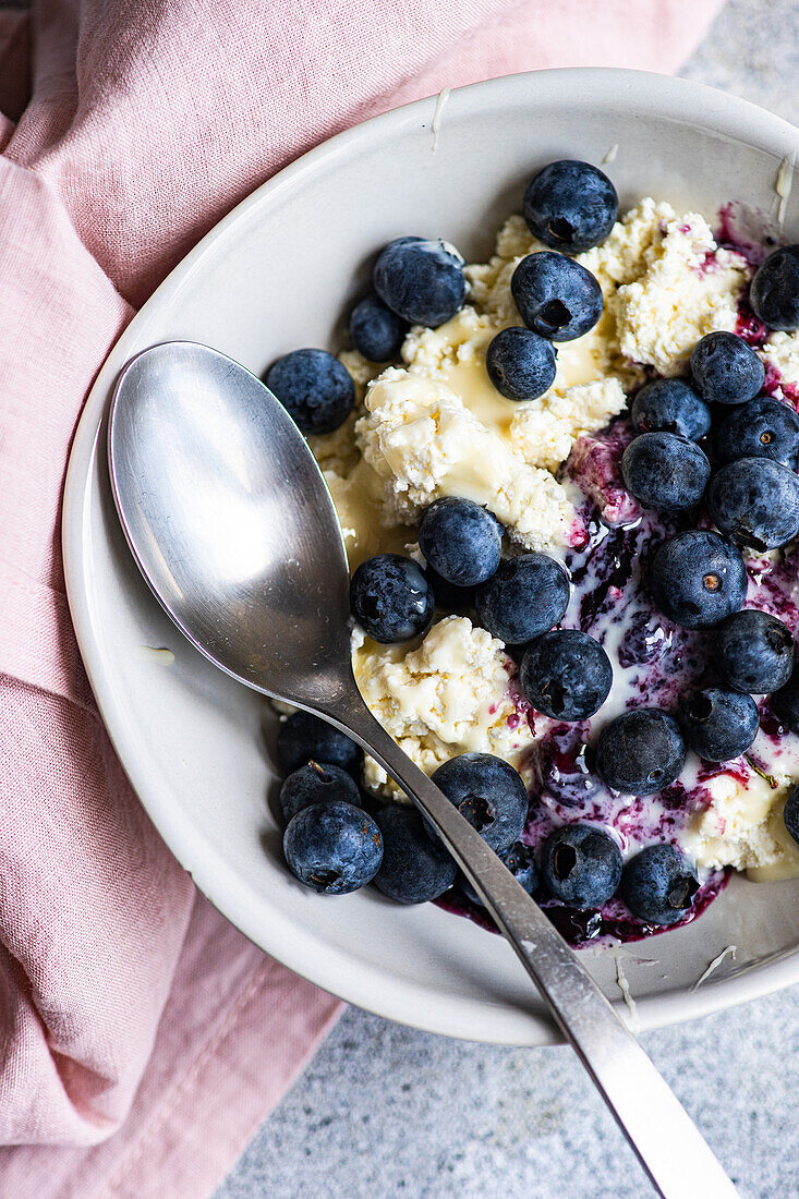 Enjoy a nutritious breakfast featuring sour cream, organic blueberry jam, and fresh blueberries atop creamy cottage cheese, all served in a tasteful ceramic bowl with a vintage spoon