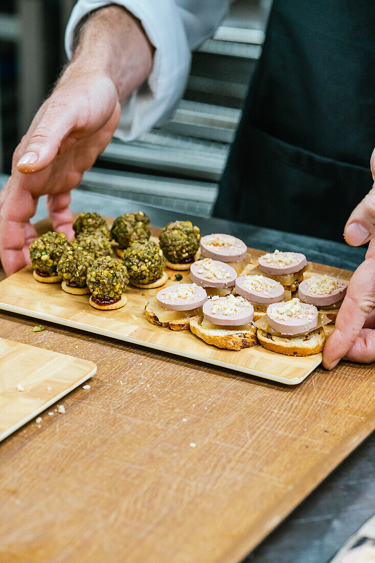 Anonymous Chef hands carefully arranging an assortment of intricate canapes topped with pâté and pistachios on a wooden serving board