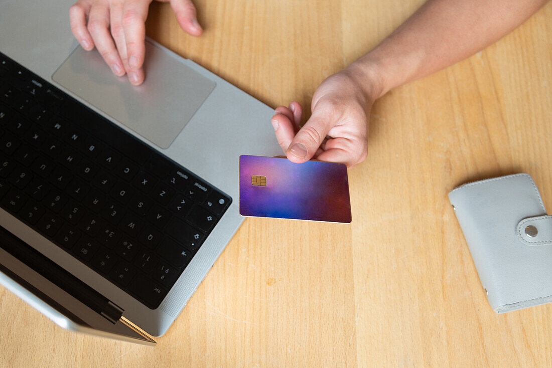 Cropped unrecognizable person hands credit card to camera while working on a laptop, showcasing easy online payment solutions at a wooden desk with a smartphone case visible.