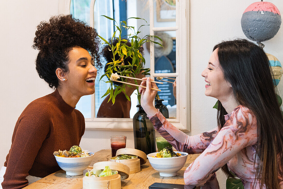 Side view of cheerful woman feeding black female friend with poke while chilling in restaurant together