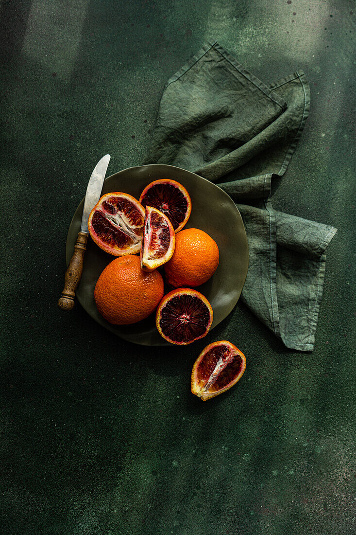 A bowl of vibrant blood oranges next to a knife on a dark, textured surface, with a moody atmosphere