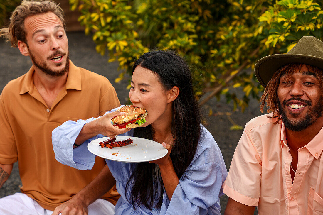 A multiethnic group of friends enjoys a BBQ outdoors. An Asian woman bites into a burger, while a Caucasian man and an African American man converse, both in casual attire and smiling.