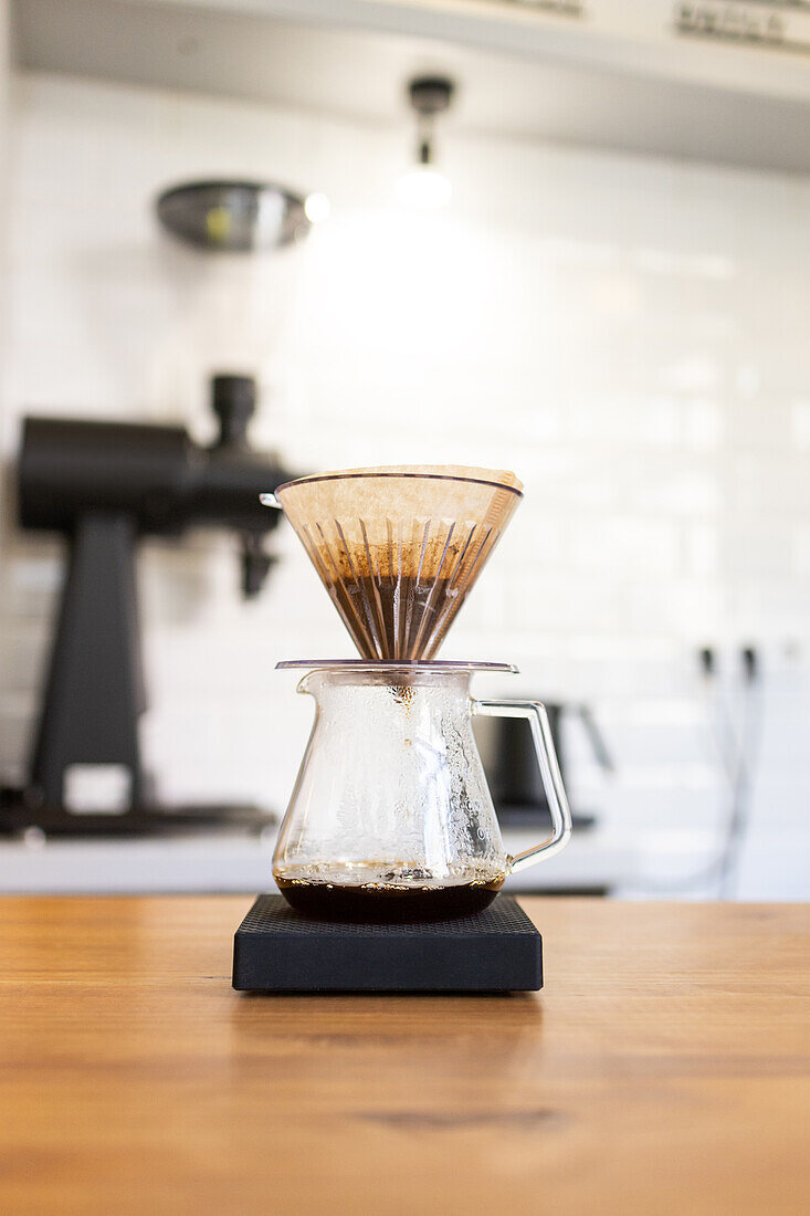 Eine Klarglaskaraffe mit einem braunen Papierfilter brüht frischen Pour-over-Kaffee auf einem Holztisch vor einer Café-Kulisse.