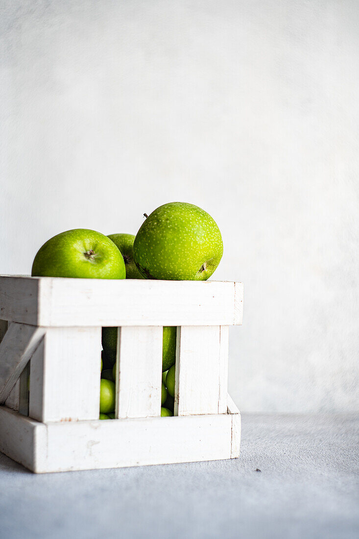 A collection of ripe green apples are neatly arranged in a white wooden crate, set against a soft gray background, invoking a fresh and organic feel