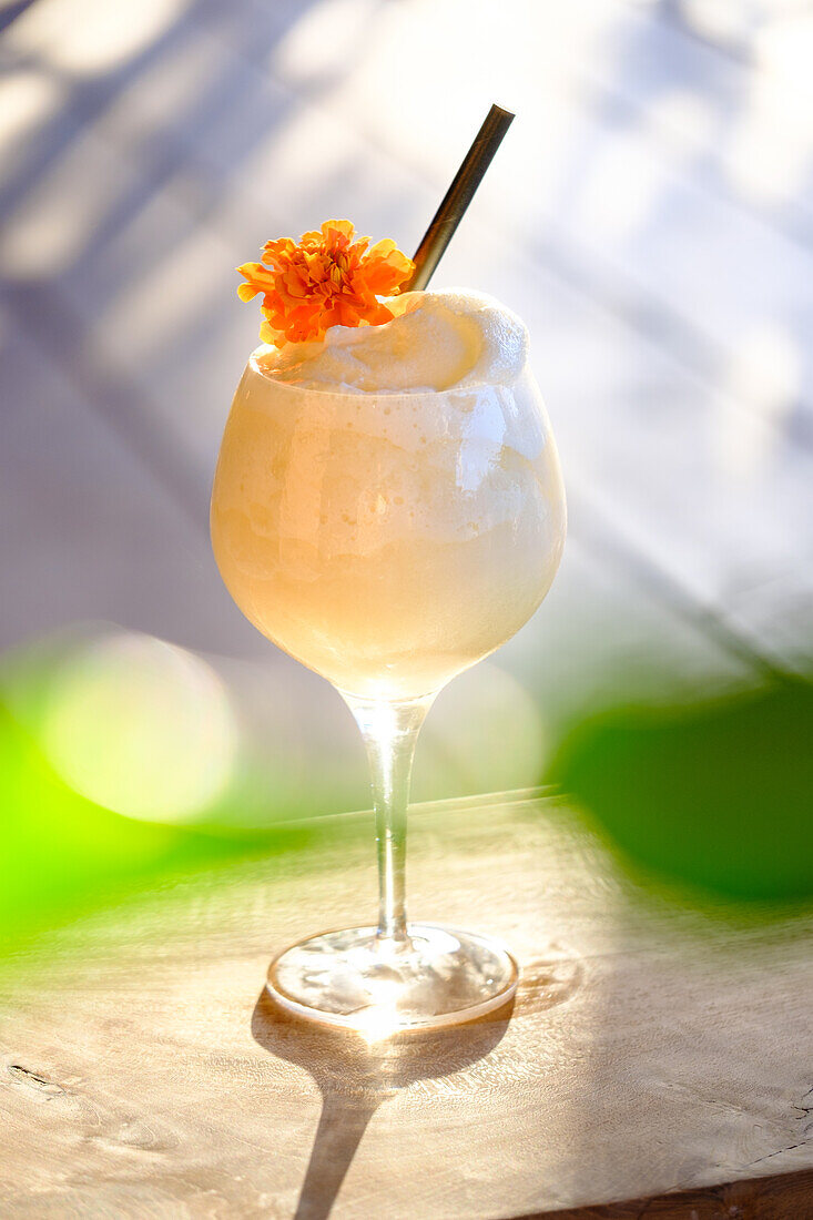 Ein erfrischender, cremiger Cocktail, wunderschön präsentiert in einem hohen Glas, gekrönt von einer leuchtend orangefarbenen Blume und einem goldenen Strohhalm, vor einem weichen, fokussierten grünen und hellen Hintergrund, der ein Gefühl von sommerlicher Eleganz vermittelt