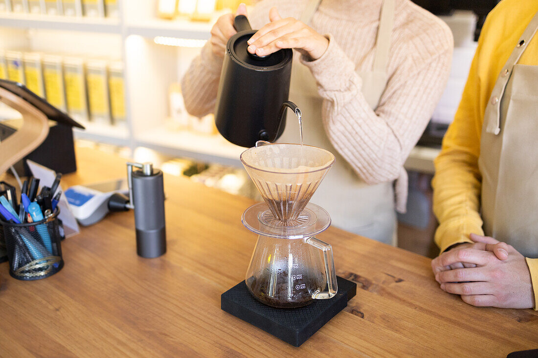 Ein Barista mit einem schwarzen Wasserkocher gießt heißes Wasser über den Kaffeesatz in einem Glastropfer, während ein Kunde wartet.