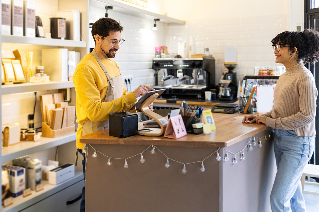 A male barista is interacting with a female customer at the counter of a well-lit, modern cafe, creating a warm and friendly atmosphere.