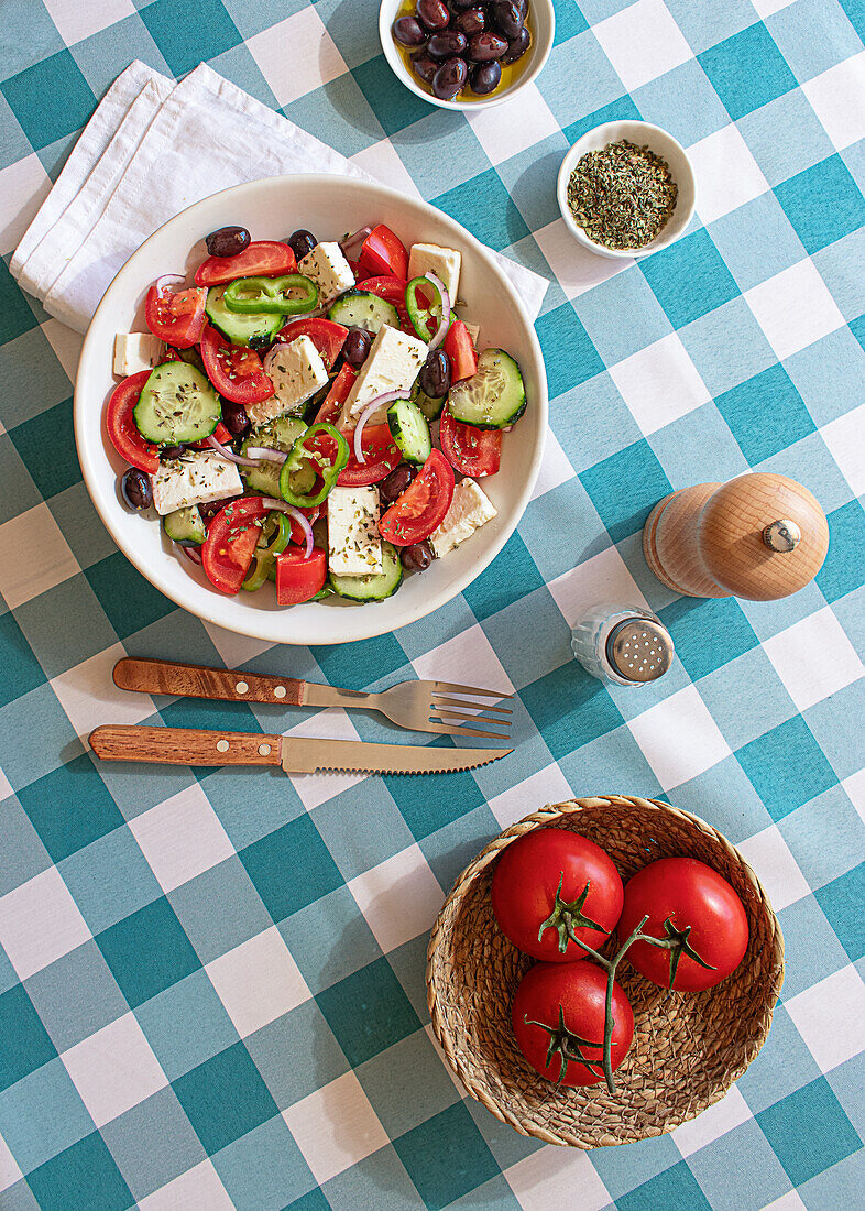 Ein bunter griechischer Salat in einer weißen Schüssel auf einer blau-weiß karierten Tischdecke, komplett mit Tomaten, Gurken, Fetakäse und Oliven, dazu ein Salzstreuer und frische Tomaten auf der Seite