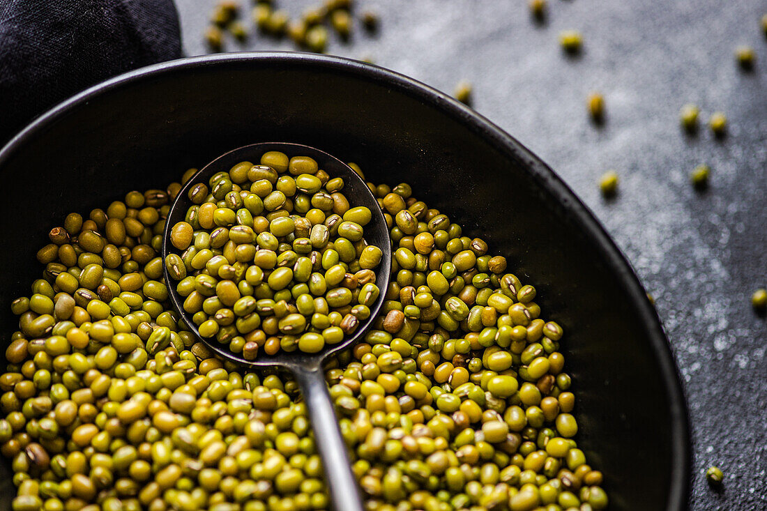 A close-up photo showcasing raw organic mung beans in a dark skillet, with a spoonful of beans amidst a scattered arrangement