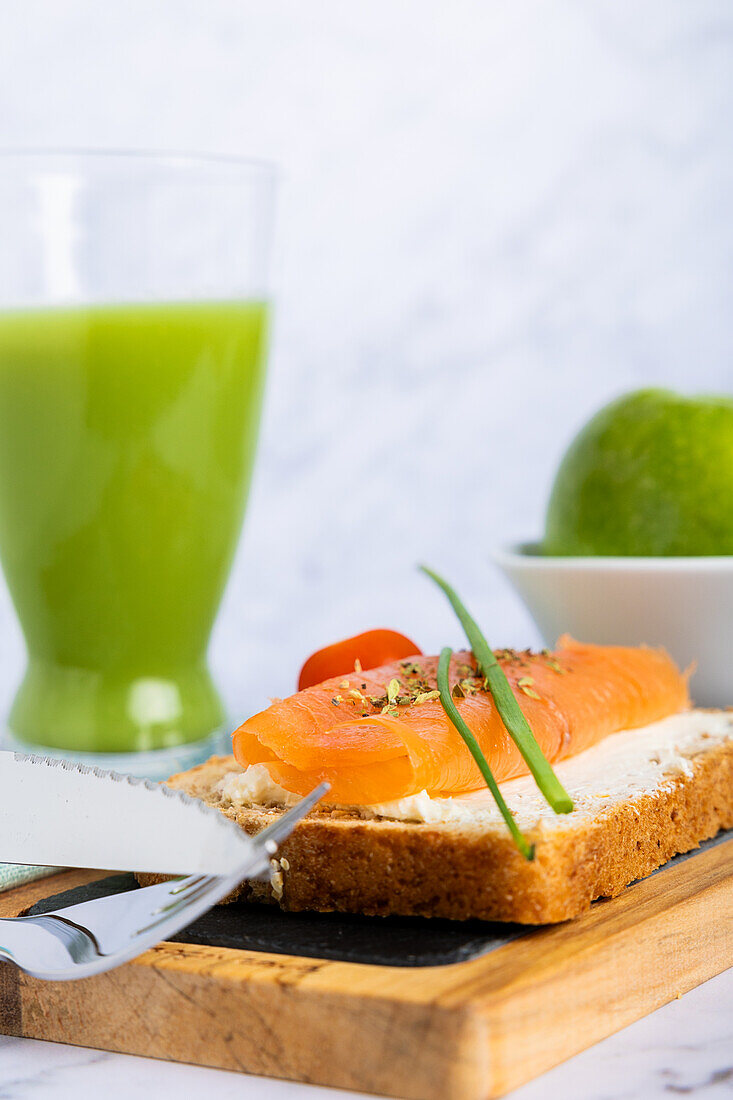 Top view of a healthy meal featuring a salmon sandwich on multigrain bread with cream cheese, topped with chives, alongside fresh cucumber and apple juice. A nourishing option for a well-balanced diet.