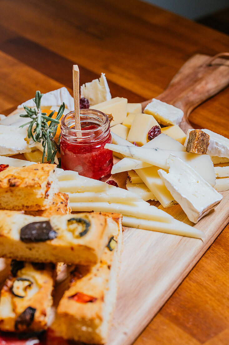 A wooden board featuring an array of artisan cheeses, slices of focaccia, and a jar of berry jam, epitomizing a gourmet snack