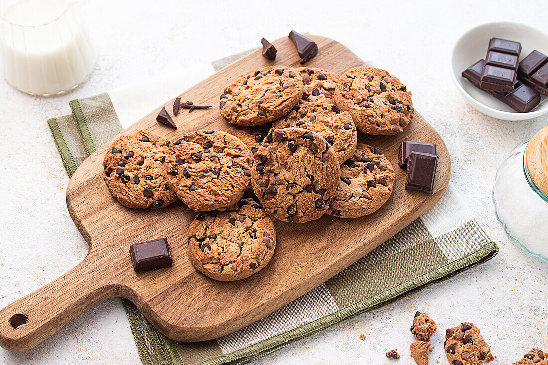 Freshly baked chocolate chip cookies on a wooden cutting board, accompanied by dark chocolate pieces and a glass of milk