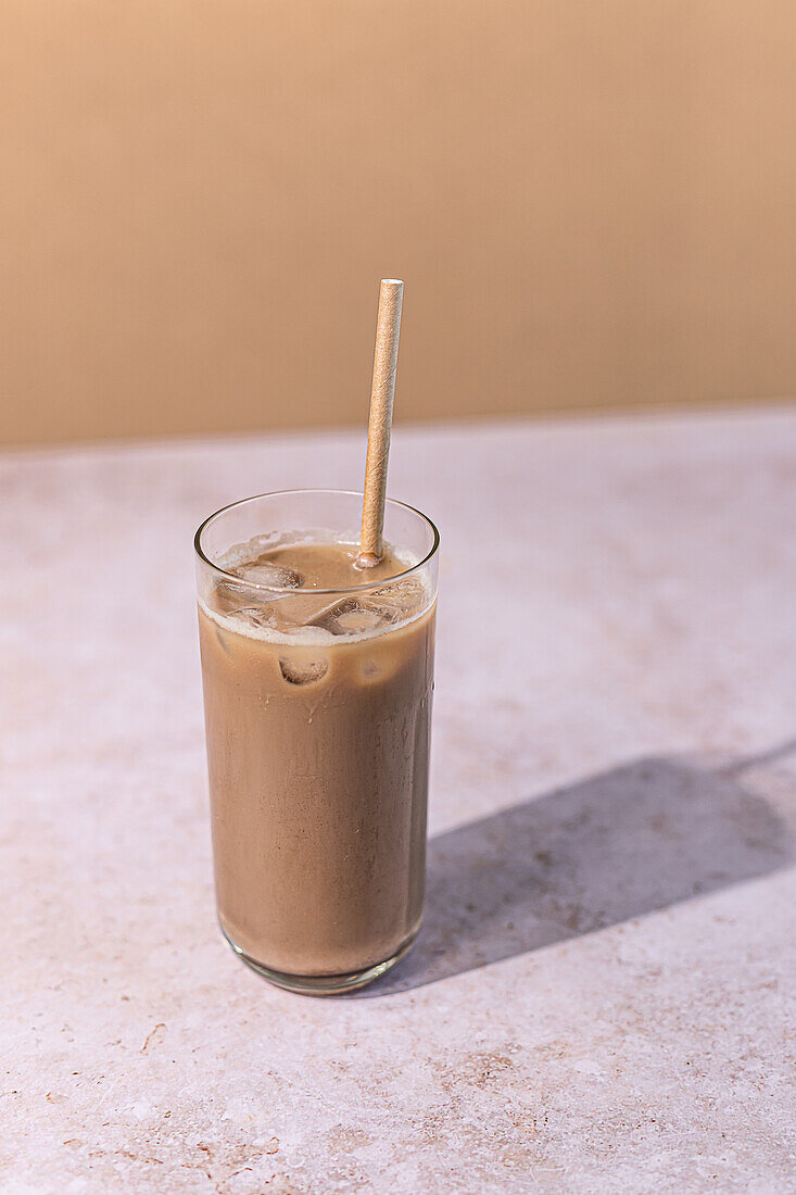 A tall glass of creamy iced coffee, complete with ice cubes and an eco-friendly straw, poised on a marble surface with a warm beige background