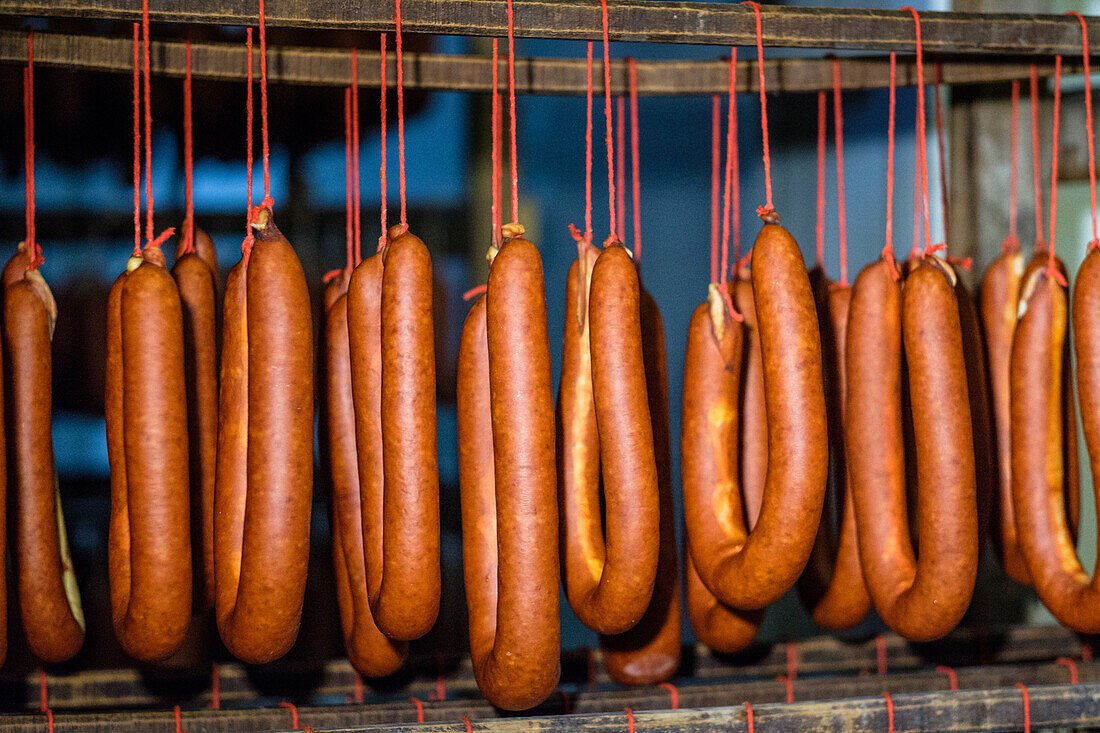 Traditionelle gepökelte Würste, darunter Sobrasada von den Balearen, hängen an roten Schnüren in einer rustikalen Räucherkammer. Die Würste sind mit Gewürzen wie Paprika gewürzt, was ihre reiche, tiefe Farbe und ihren Geschmack hervorhebt.