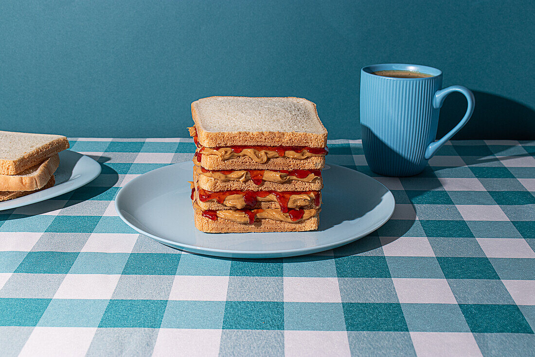 Ein köstliches Erdnussbutter-Gelee-Sandwich wird auf einem blauen Teller, einer Tasse amerikanischen Kaffees und einigen Brotscheiben vor einem blauen Hintergrund serviert