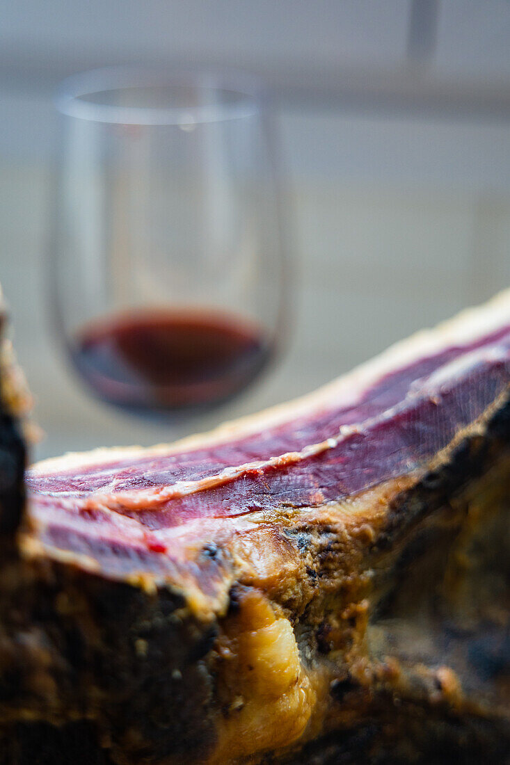 Diese Nahaufnahme zeigt ein köstliches Stück gepökelten iberischen Schinken mit einem Glas Rotwein im Hintergrund Die satten Farben und Texturen unterstreichen die Eleganz und den Geschmack traditioneller Gourmet-Paarungen