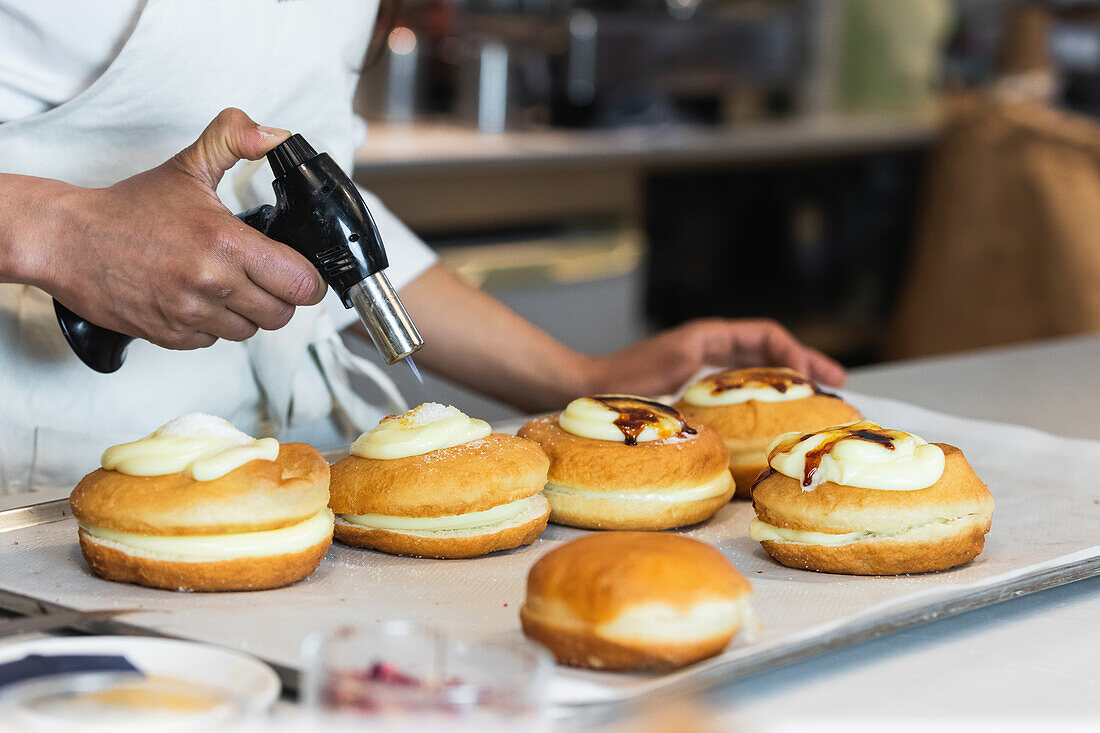 Unrecognizable crop chef caramelizing sugar cream on sweet Berliners with gas torch while preparing vegan dessert in bakehouse