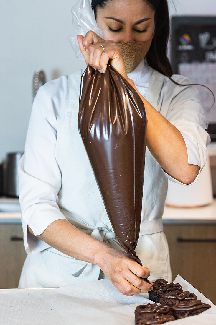 Eine Bäckerin mit Maske drückt Schokoladencreme aus einem Spritzbeutel auf leckere vegane Biskuitkuchen, während sie in der Küche einer Bäckerei Desserts zubereitet