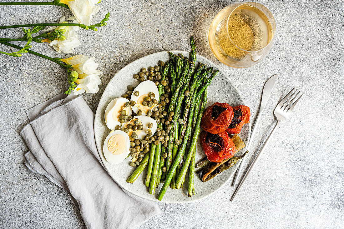 Top view of nutritious lunch setup featuring boiled eggs, barbecued asparagus, capers, and grilled vegetables including tomatoes and chili peppers, presented elegantly on a speckled plate with a glass of wine and cutlery.