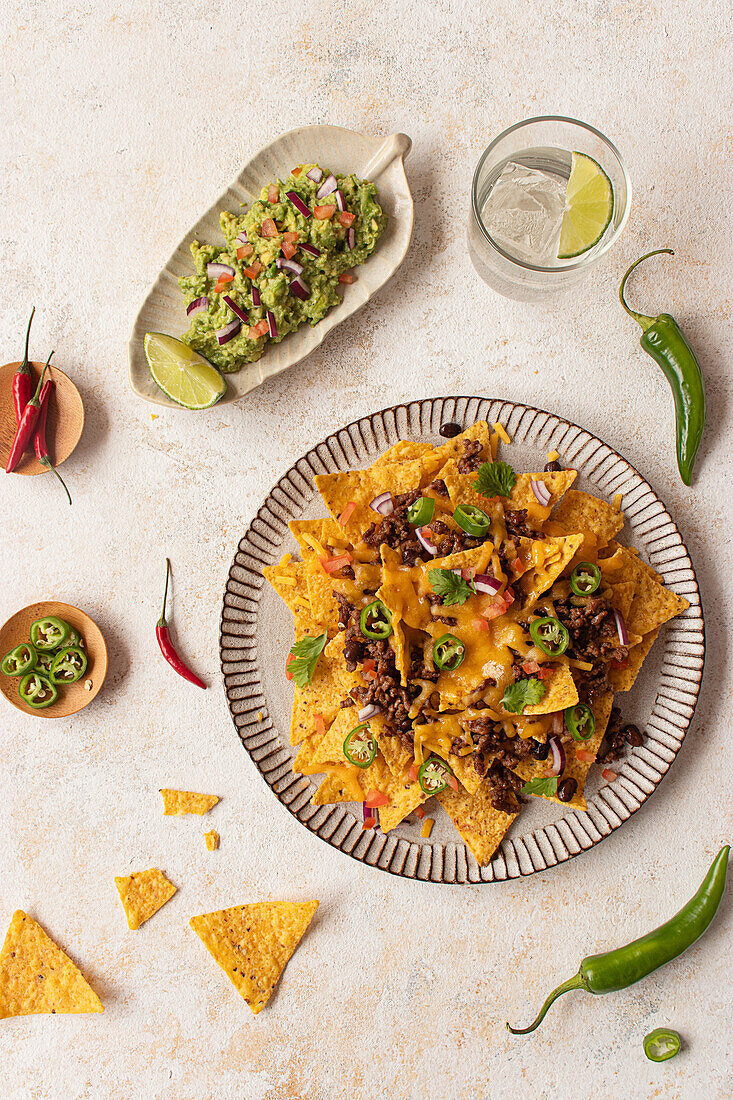 Top view of a plate of nachos generously topped with cheese, ground meat, jalapenos, and fresh herbs, served alongside a dish of guacamole and a refreshing lime drink on a rustic background.