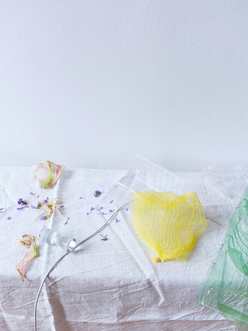 A minimalist artistic arrangement featuring dried flower petals scattered around a vivid yellow net, placed delicately on a textured white fabric, accented by soft purple petals and subtle lighting