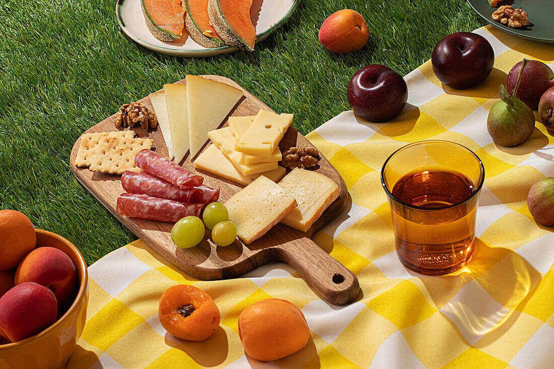 An outdoor summer picnic spread featuring a variety of cheeses, cured meats, and fresh fruits on a yellow and white checkered blanket