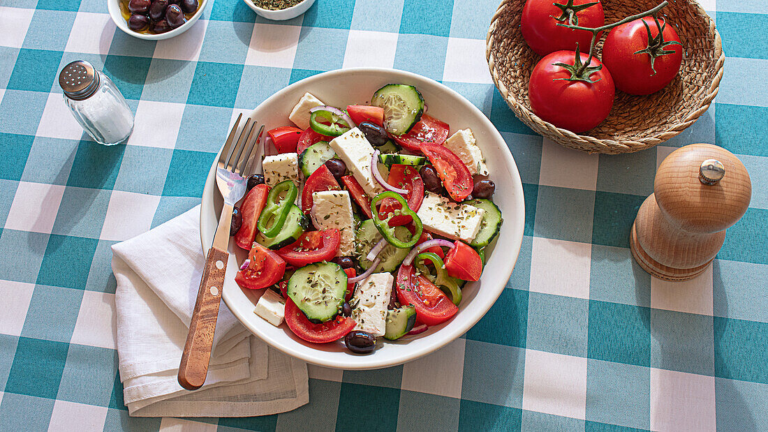 Colorful Greek salad with tomatoes, cucumbers, onions, feta cheese, and olives served on a white bowl, set on a checkered blue and white tablecloth